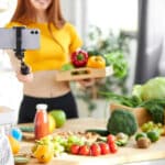 Ein Food-Blogger hält frische Lebensmittel in der einen Hand, in der anderen Hand hält sie einen Selfie-Stick Fokus auf das Handy. © Roman - stock.adobe.com