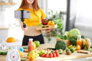 Ein Food-Blogger hält frische Lebensmittel in der einen Hand, in der anderen Hand hält sie einen Selfie-Stick Fokus auf das Handy. © Roman - stock.adobe.com