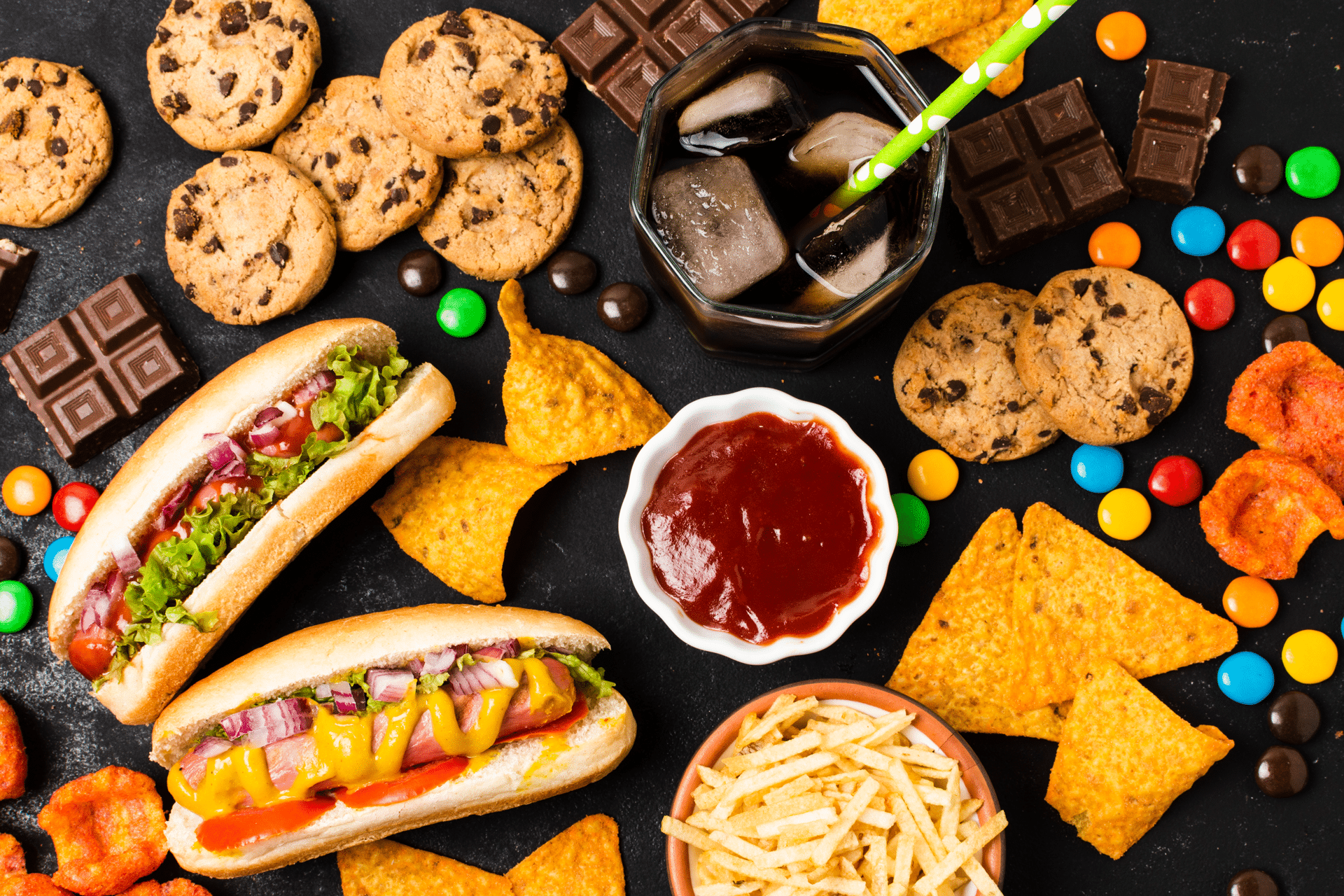 Hot Dogs, Pommes, Kekse, Schokolade, Schokolinsen, Ketchup, Tacos und ein eisgefühlte Limonade mit Strohhalm stehen auf einem Tisch. Image by freepik