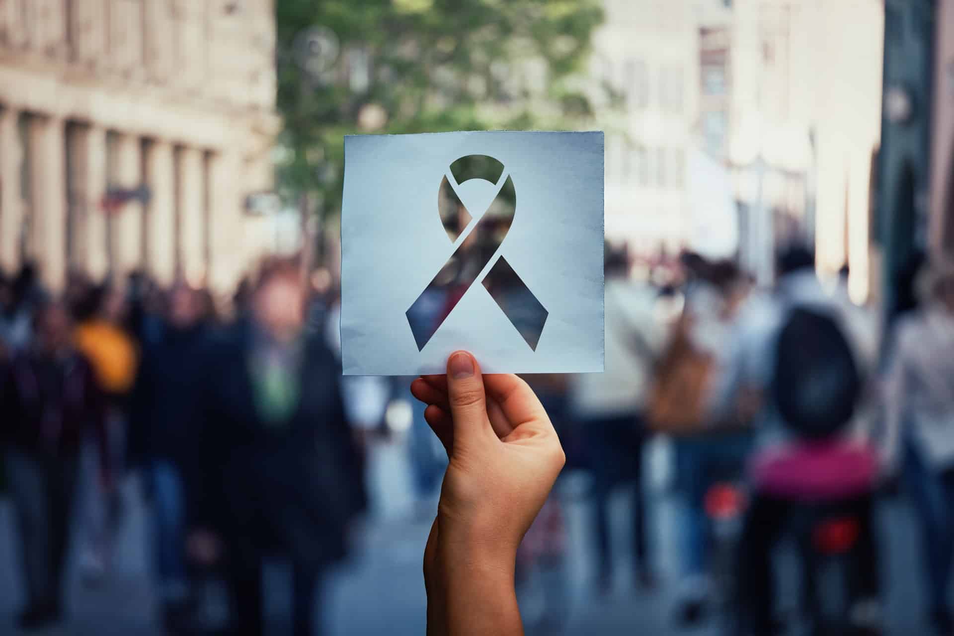 Hand hält Menschen in der Öffentlichkeit ein Blatt Papier mit einer Aidsschleife hin. © 1STunningART - stock.adobe.com